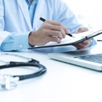 Doctor working with laptop computer and writing on paperwork. Hospital background.
